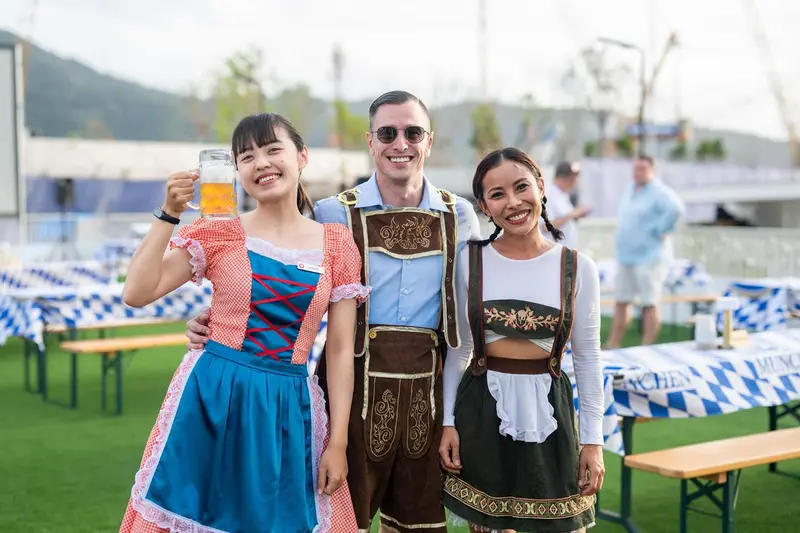 Trois personnes souriantes, en tenue traditionnelle, tiennent des chopes de bière sur une pelouse.