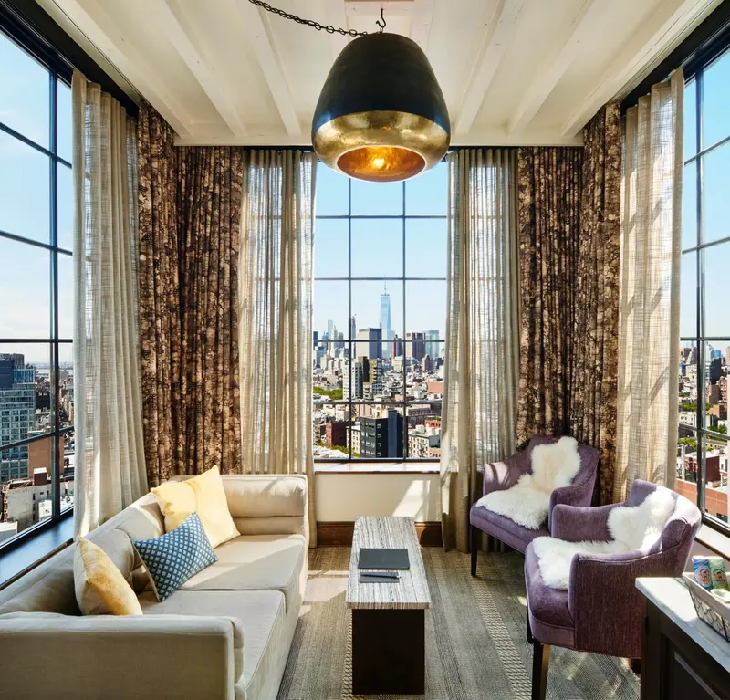 Un salon lumineux avec canapés et fauteuils face à une grande fenêtre offrant une vue sur la ville.