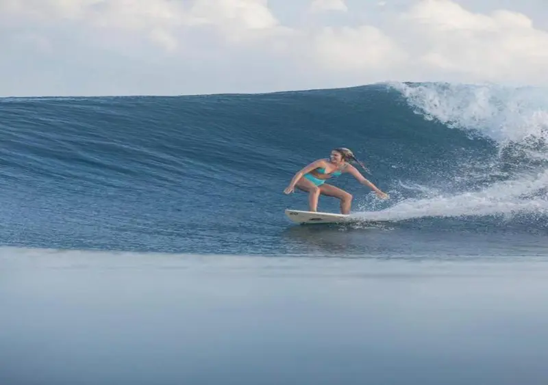 Une surfeuse chevauche une vague turquoise sous un ciel nuageux.