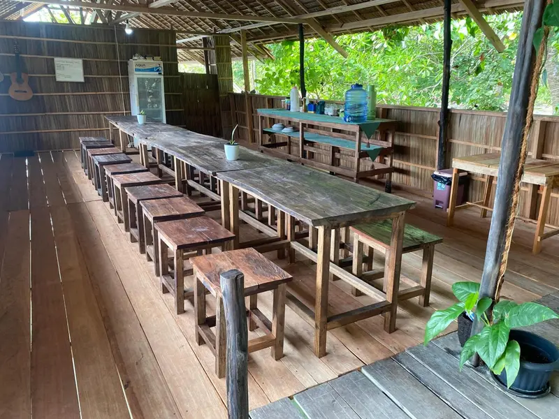 Une longue table en bois est entourée de bancs dans un espace ouvert et ombragé.