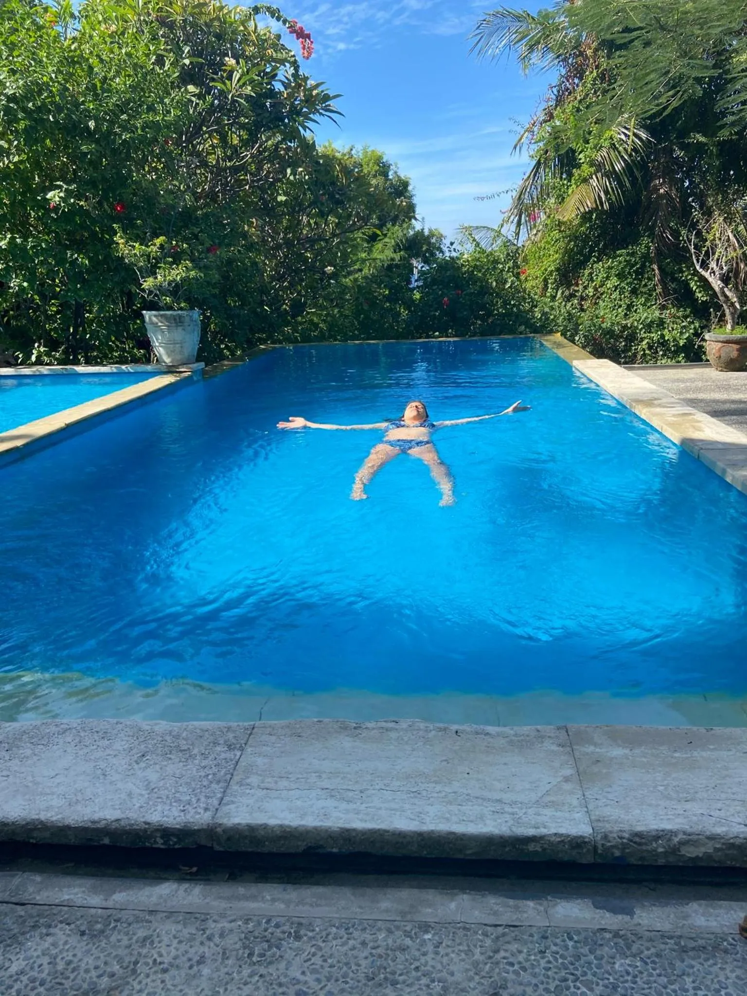 Une personne flotte sur le dos dans une piscine rectangulaire bordée d'une terrasse.