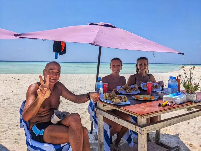 Trois personnes sourient et font le signe de la victoire autour d'une table de pique-nique sur une plage ensoleillée.