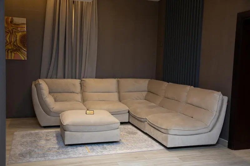 Un canapé d'angle beige et un repose-pieds sont disposés sur un tapis dans un intérieur clair.