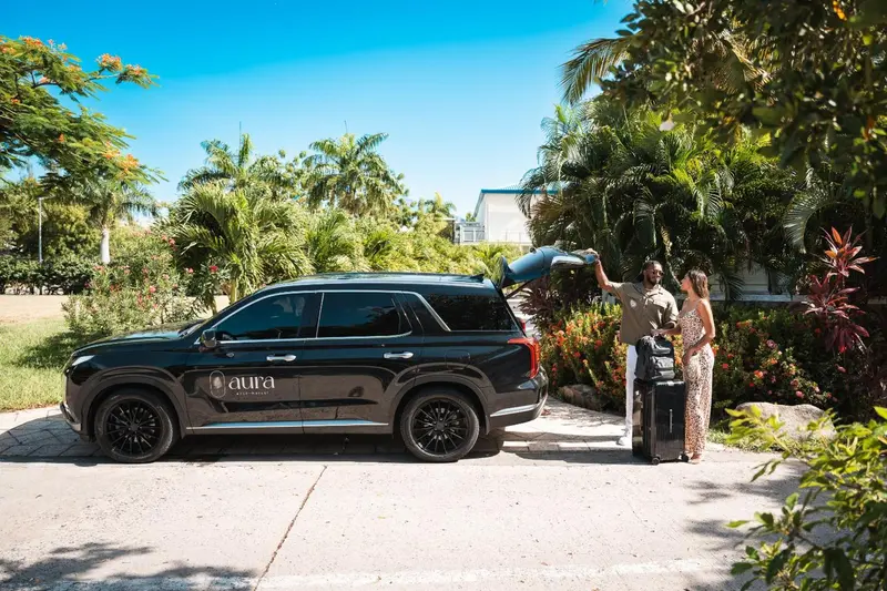 Une voiture noire est garée sur un chemin bordé de végétation tropicale, avec des passagers déchargeant des bagages.