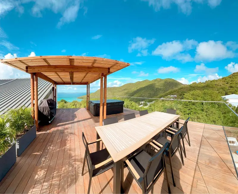 Une table à manger en bois est placée sur une terrasse ensoleillée avec vue sur un paysage verdoyant.
