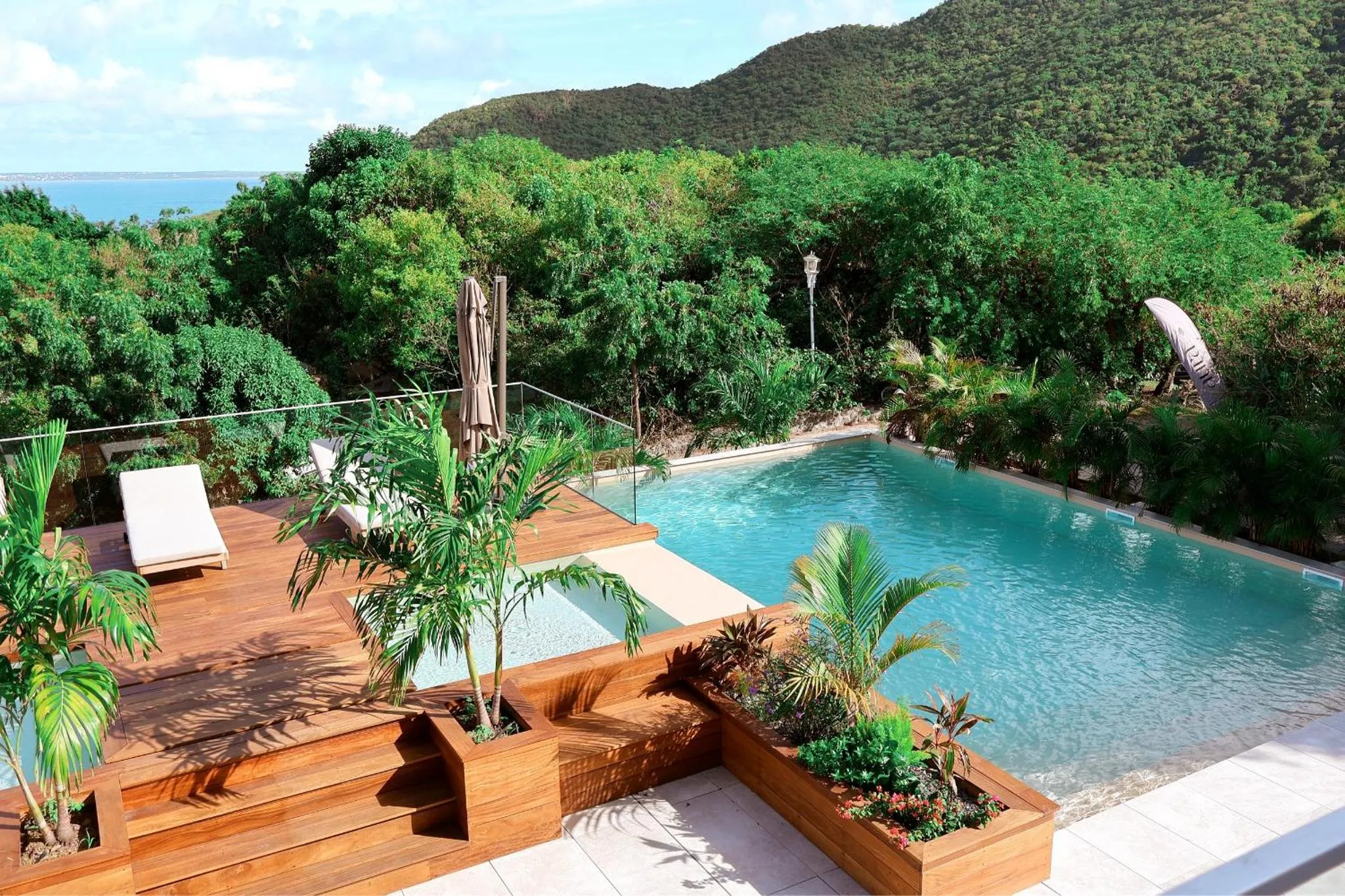 Terrasse en bois avec transats surplombant une piscine turquoise et une vue sur la mer.