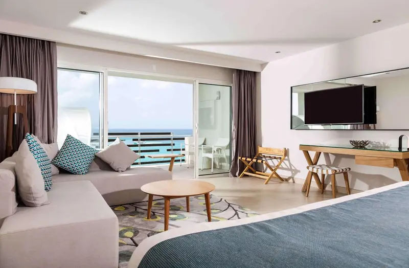 Une chambre lumineuse avec un canapé, un bureau et un accès à un balcon avec vue sur la mer.