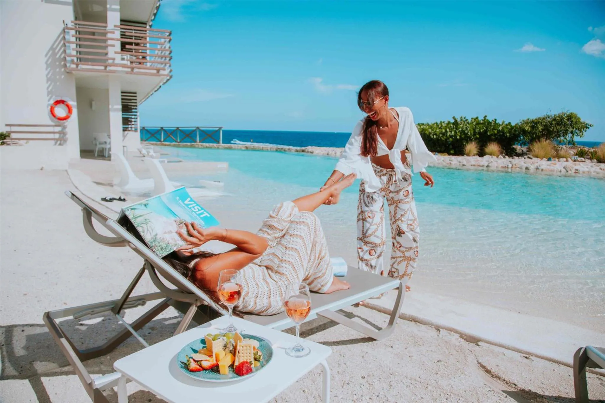 Deux femmes se détendent au bord d'une piscine avec des boissons et des fruits.