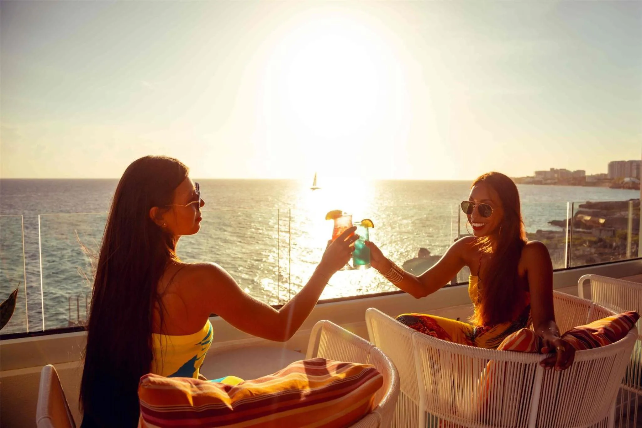 Deux femmes trinquent avec des cocktails sur une terrasse ensoleillée face à la mer.