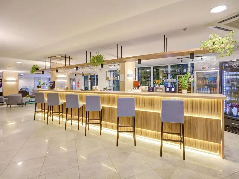 Un long comptoir de bar en bois clair est éclairé d'une lumière chaleureuse, avec des tabourets hauts et des bouteilles exposées.