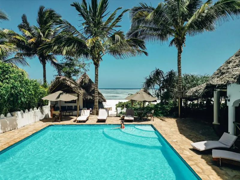 Une piscine turquoise bordée de transats et de palmiers, avec une plage en arrière-plan.