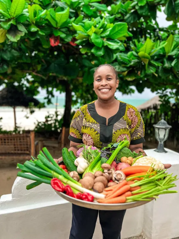 Une femme souriante présente un panier rempli de légumes frais devant un décor tropical.