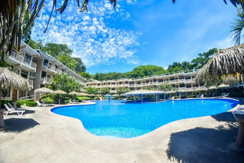 Une piscine turquoise bordée de chaises longues et de palmiers, devant un bâtiment à plusieurs étages.