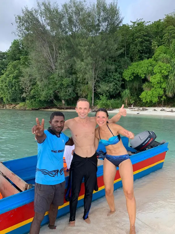 Trois personnes souriantes posent sur un bateau coloré près d'une plage.