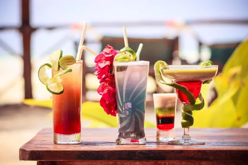 Trois cocktails colorés ornés de fruits et de fleurs sont présentés sur une table en bois.