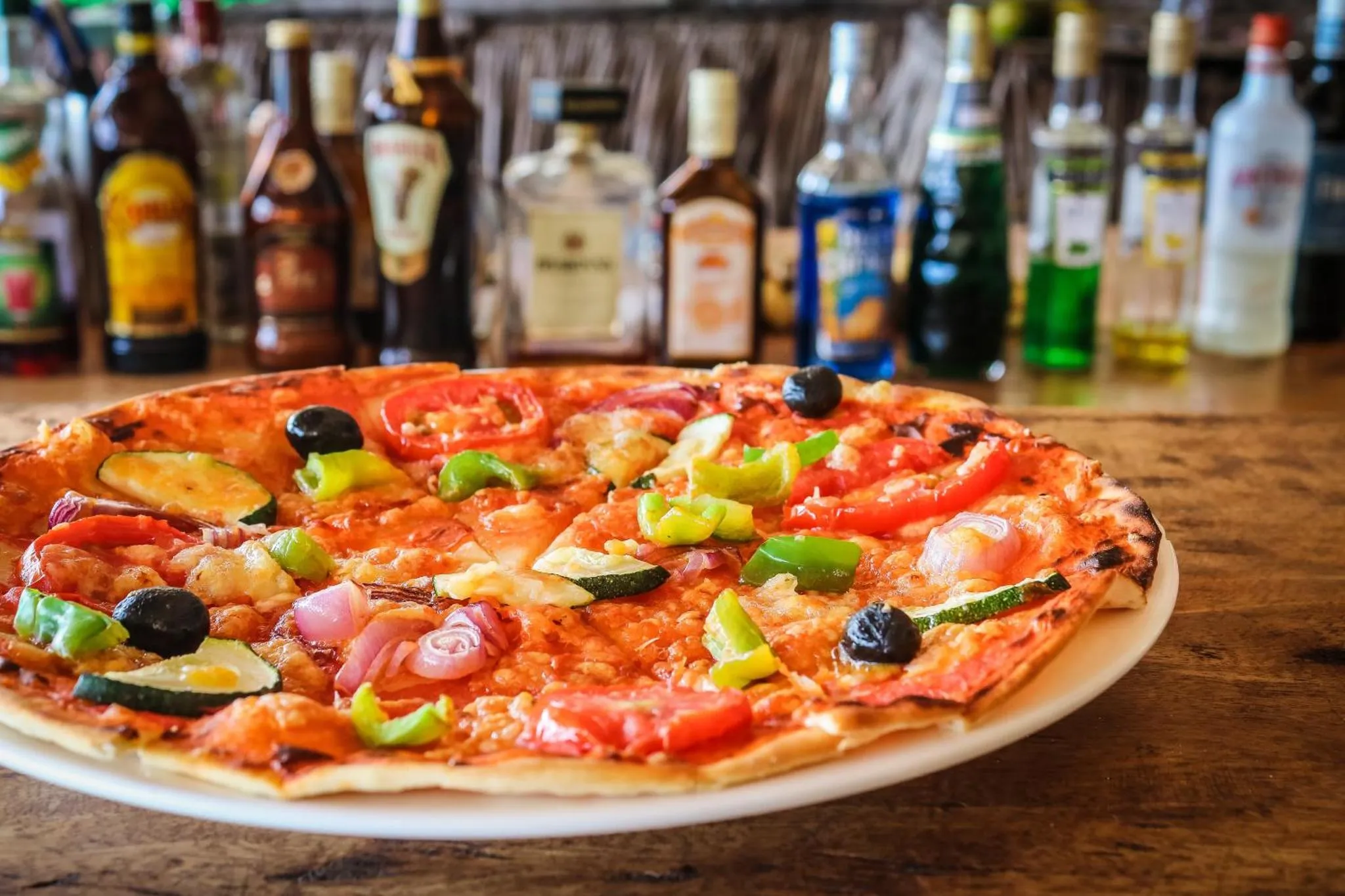 Une pizza colorée est présentée sur un comptoir de bar avec des bouteilles en arrière-plan.