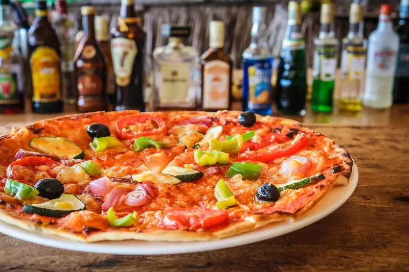 Une pizza colorée est présentée sur un comptoir de bar avec des bouteilles en arrière-plan.