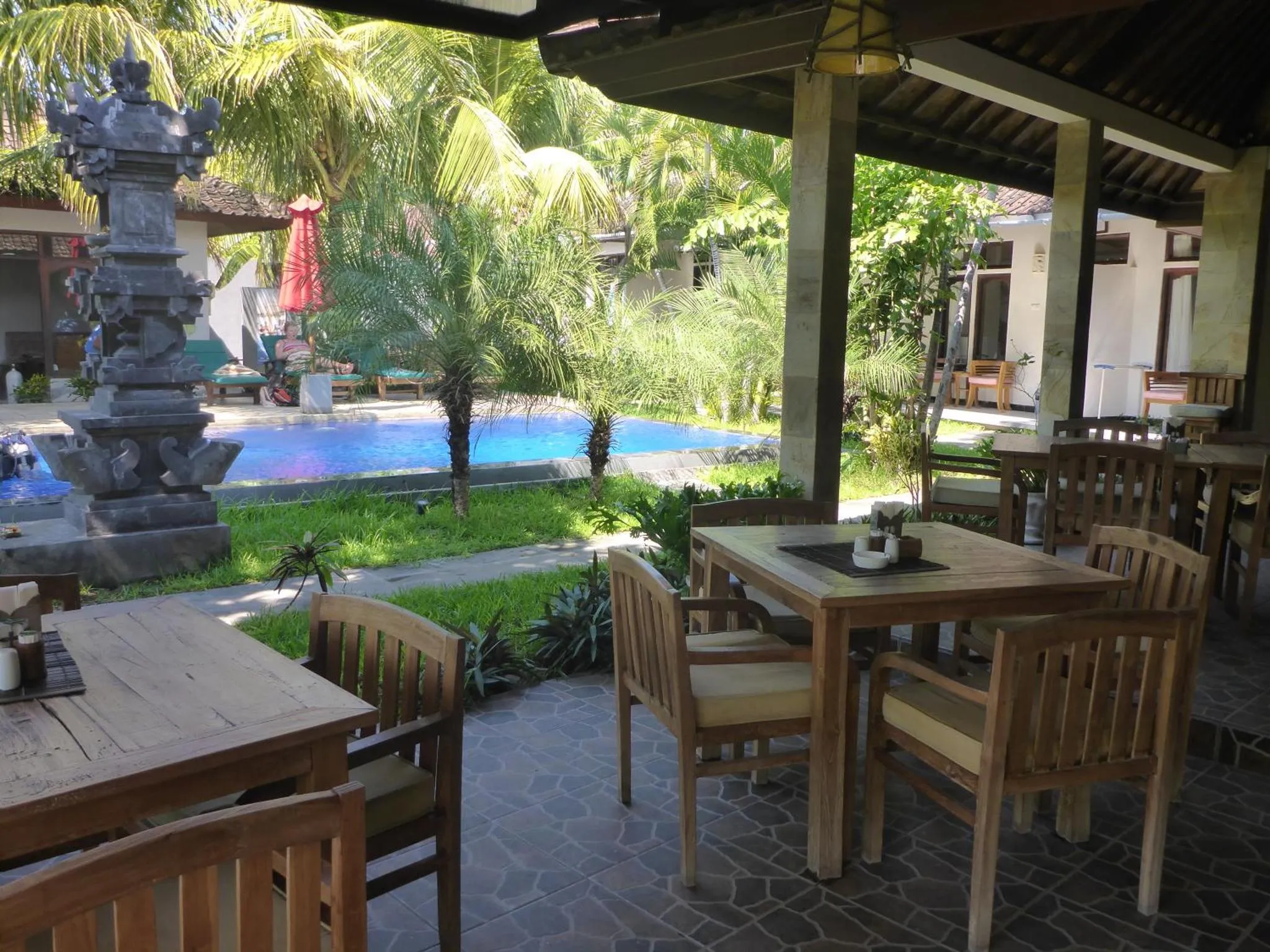 Tables et chaises en bois disposées sur une terrasse ombragée près d'une piscine.