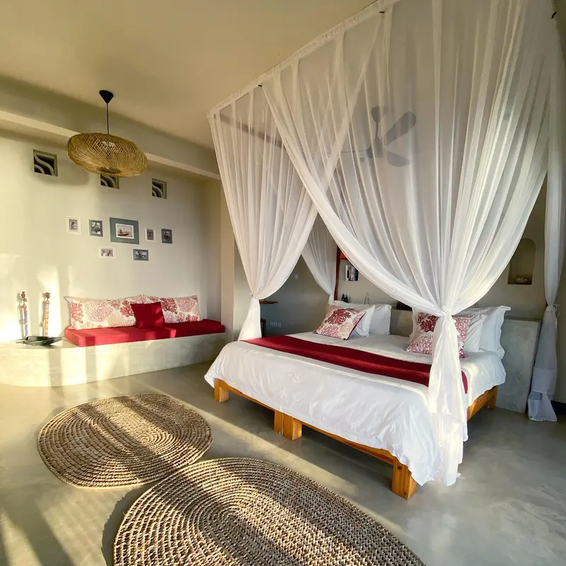 Une chambre lumineuse avec un lit à baldaquin et une banquette rouge.