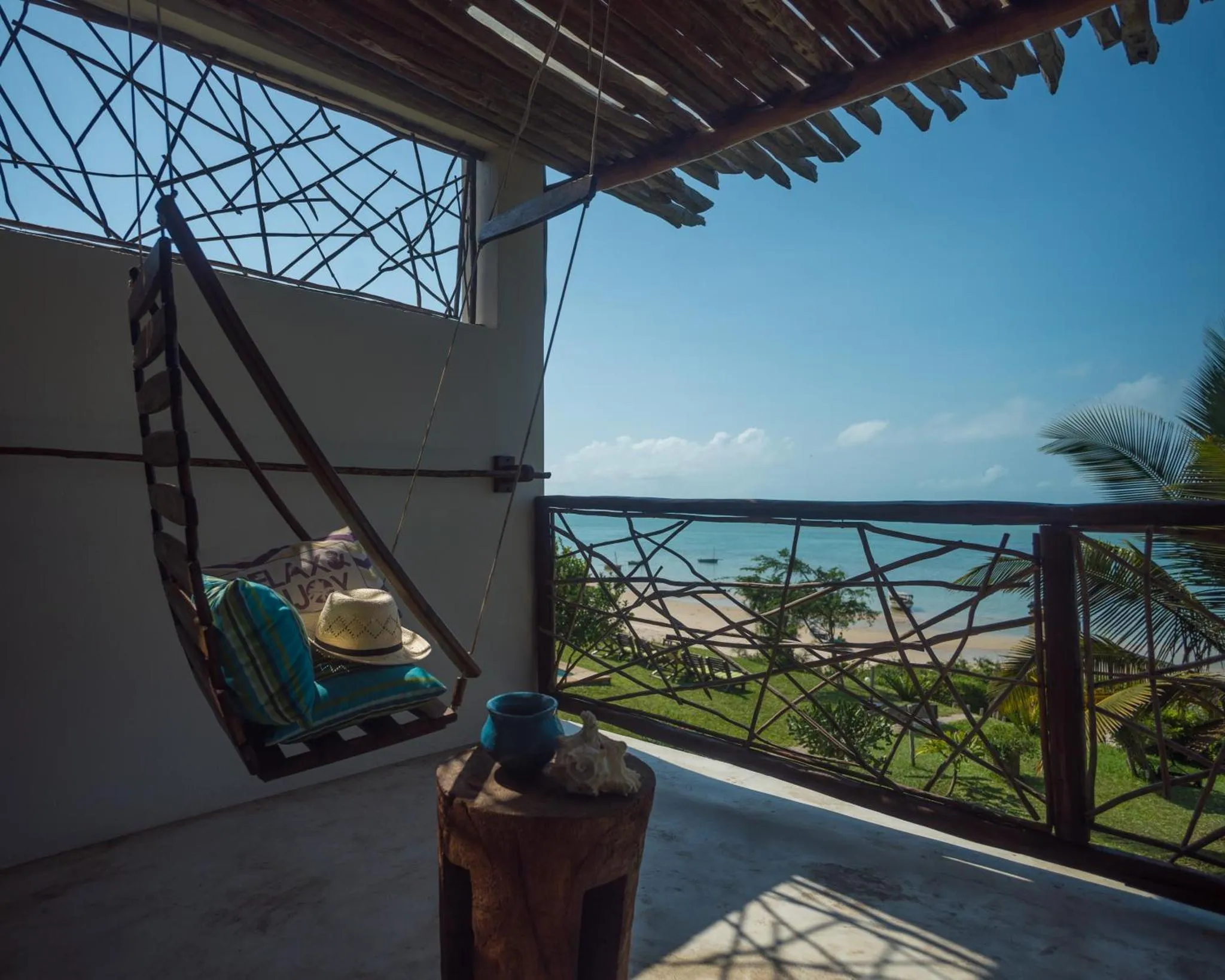 Un hamac bleu et vert est suspendu sur une terrasse ensoleillée avec vue sur la mer.