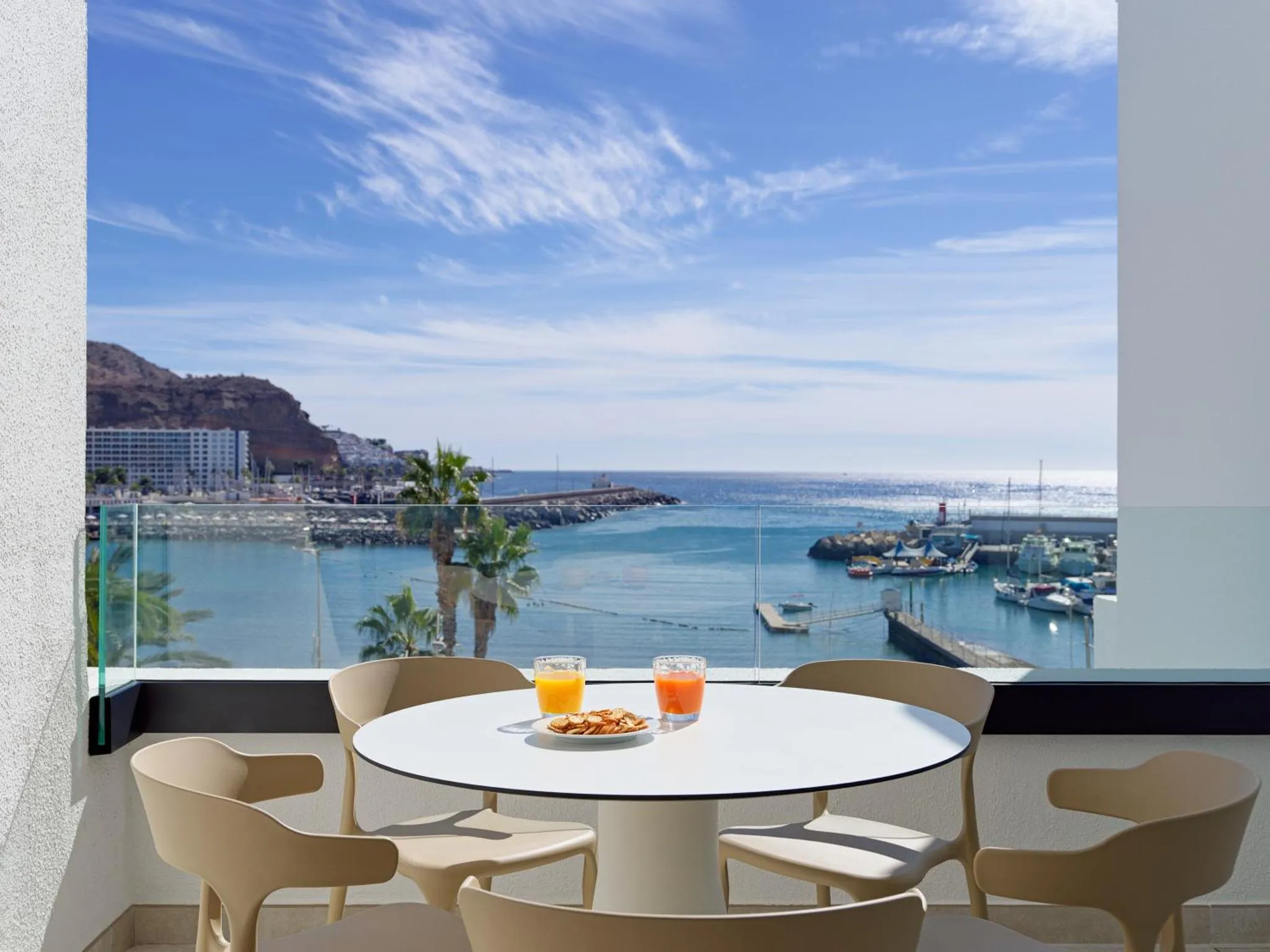 Une table ronde avec des boissons et des en-cas sur une terrasse ensoleillée avec vue sur la mer et un port.