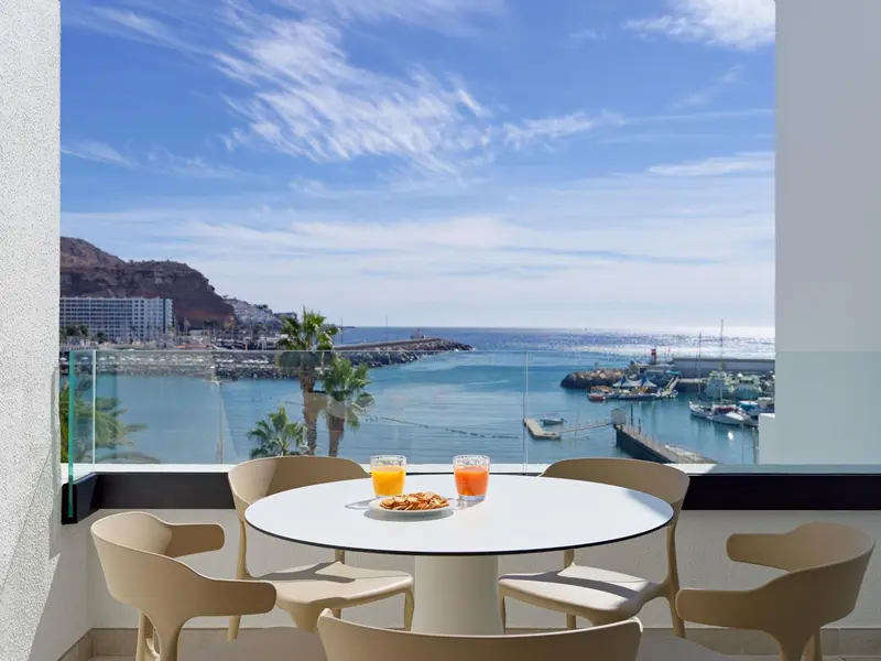 Une table ronde avec des boissons et des en-cas sur une terrasse ensoleillée avec vue sur la mer et un port.