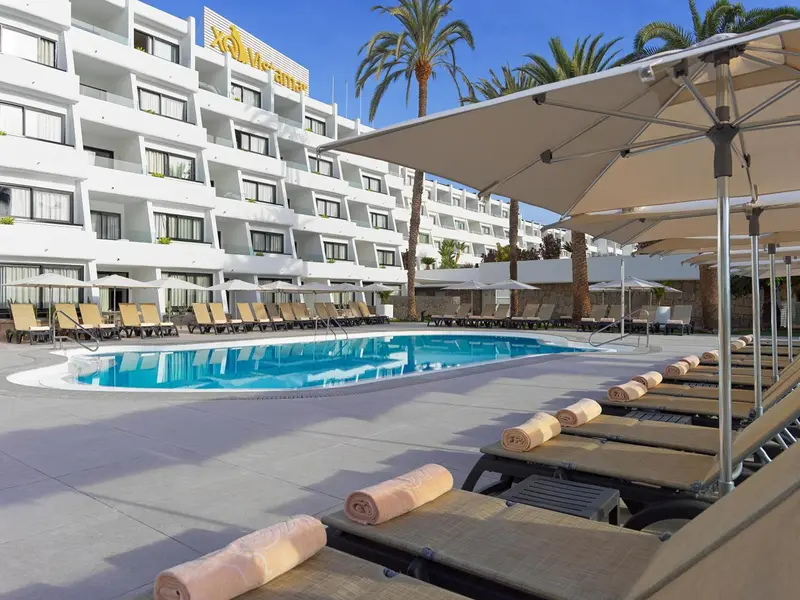 Transats alignés au bord d'une piscine ensoleillée devant un bâtiment blanc.