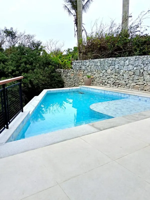 Une piscine rectangulaire avec un bord arrondi est entourée d'une terrasse carrelée et d'une végétation luxuriante.