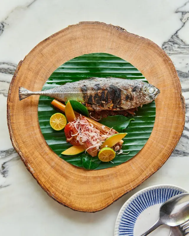 Un poisson grillé est présenté sur une assiette en bois avec des accompagnements colorés.