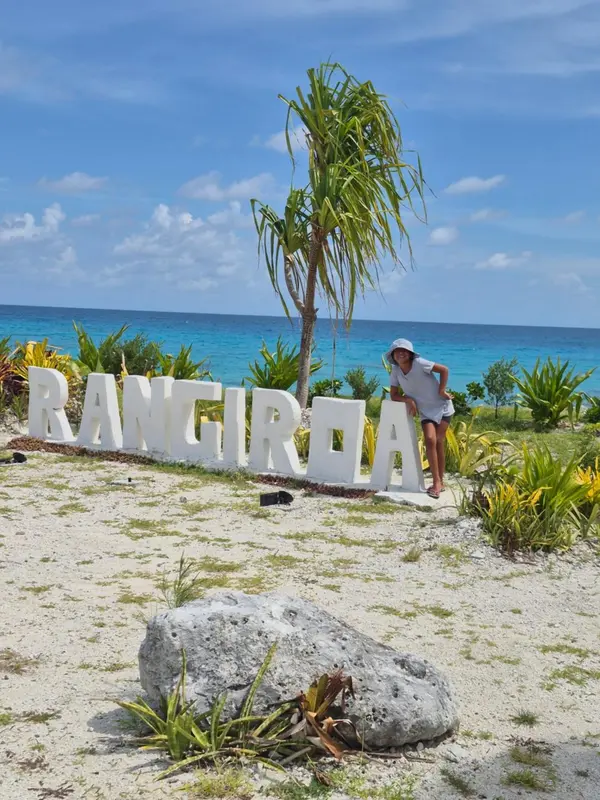 Une personne se tient devant de grandes lettres blanches sur une plage de sable clair.