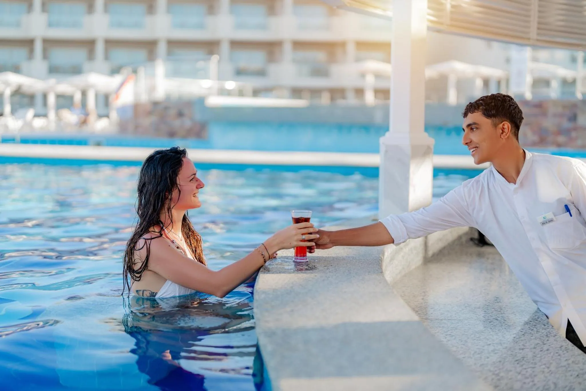 Un couple souriant échange un verre au bord d'une piscine ensoleillée.