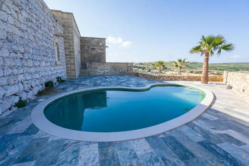 Une piscine rectangulaire avec un bord arrondi est entourée d'une terrasse pavée et de murs en pierre.