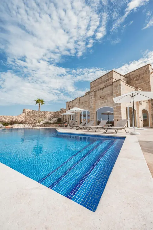 Piscine turquoise bordée d'une terrasse ensoleillée devant un bâtiment clair.