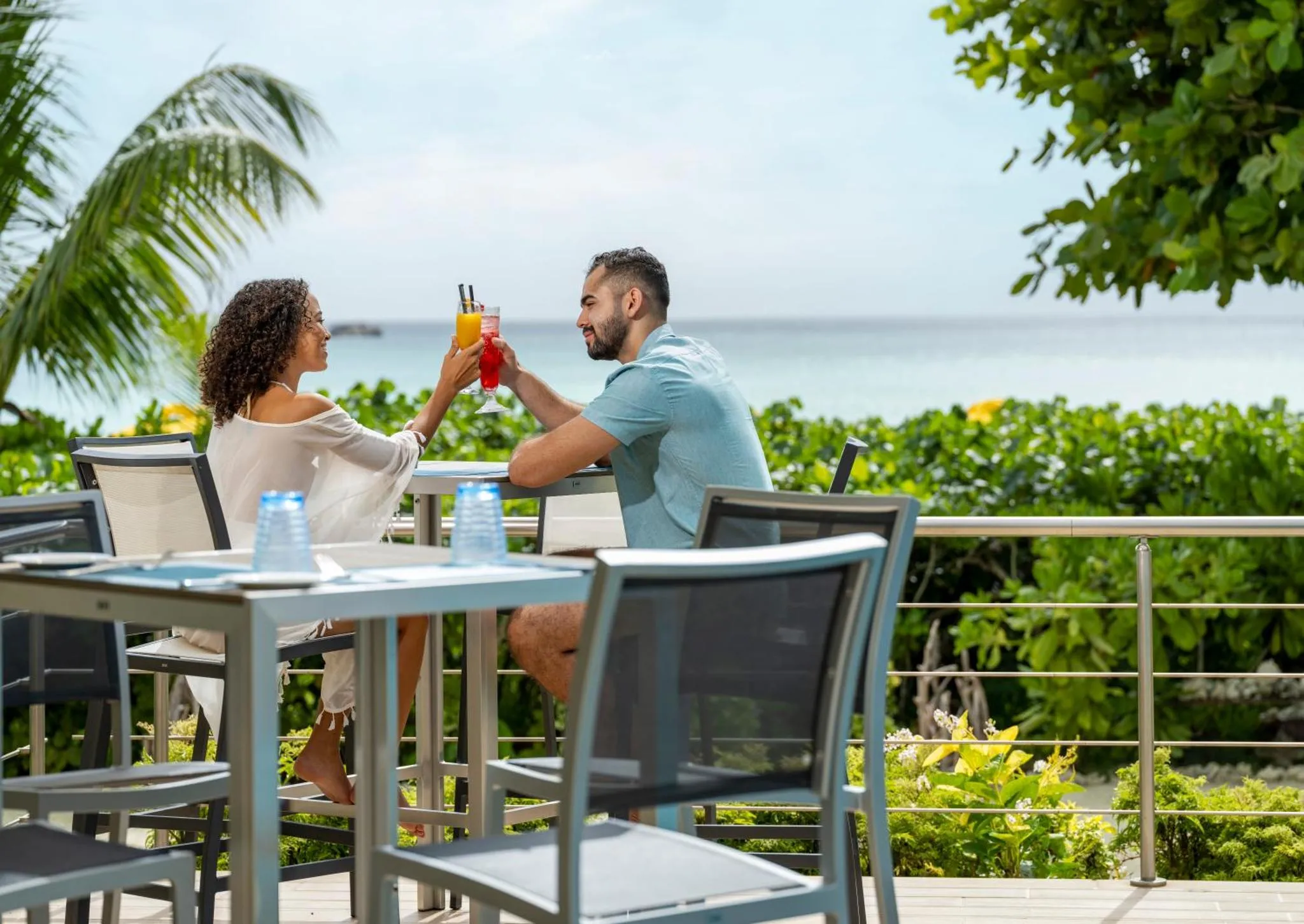Un couple trinque avec des cocktails sur une terrasse ensoleillée face à la mer.