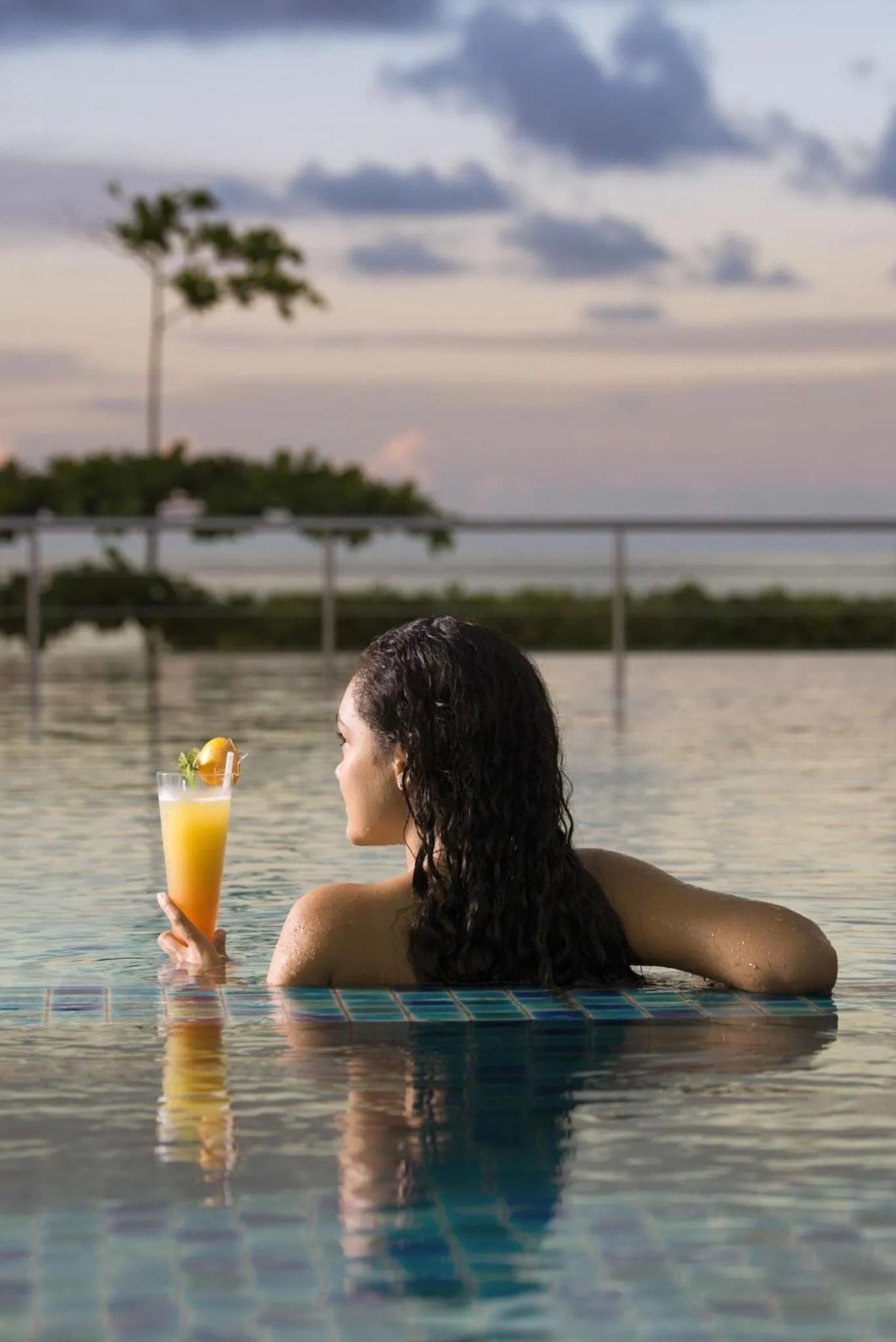 Une femme se détend dans une piscine avec un cocktail à la main, face à la mer.