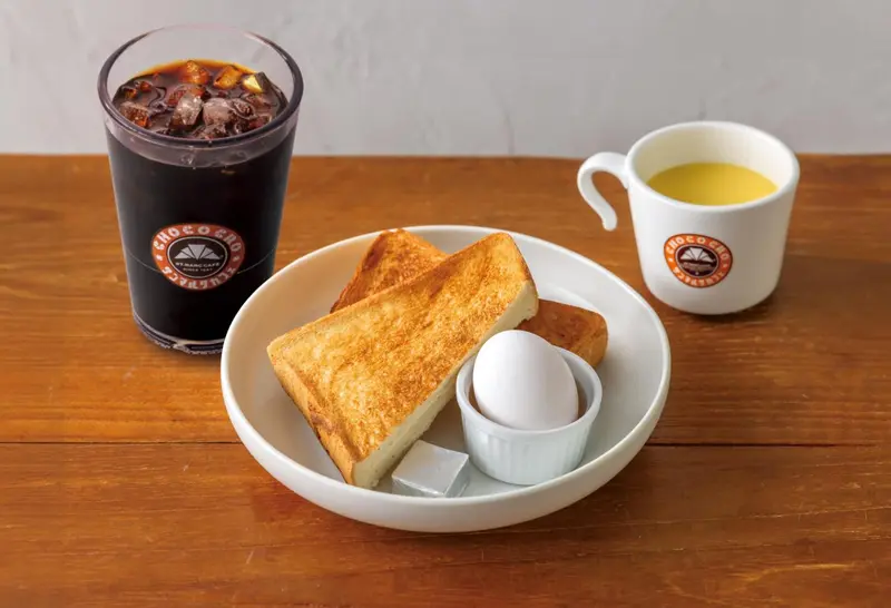 Un petit-déjeuner avec une boisson sombre, des toasts, un œuf et une tasse de liquide jaune.