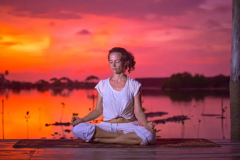 Une femme est assise en tailleur sur une plateforme en bois face à l'eau au coucher du soleil.