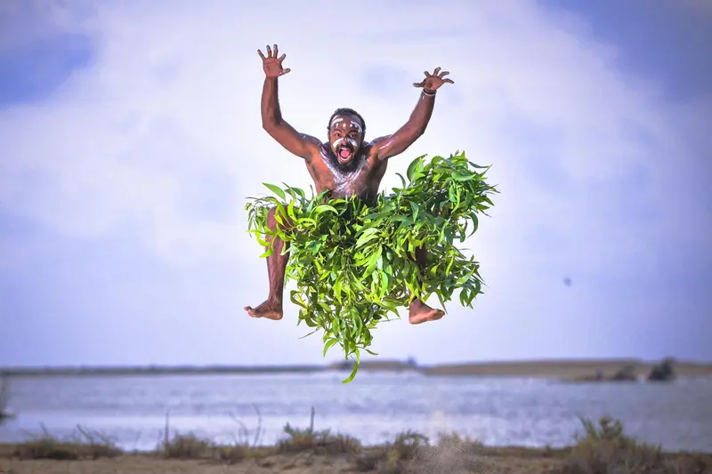 Un homme saute en l'air, recouvert de feuillage vert sur une plage.