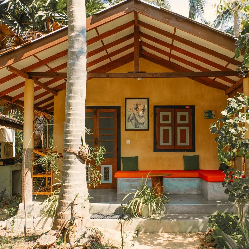 Une petite maison jaune avec une terrasse ombragée et une végétation luxuriante.