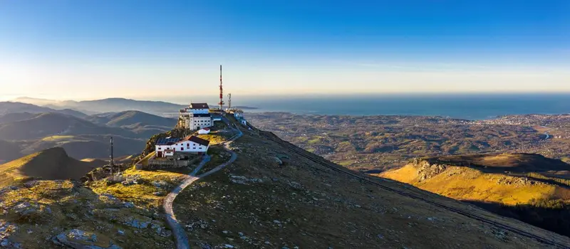 Vue aérienne d'un bâtiment perché sur une montagne verdoyante avec la mer à l'horizon.