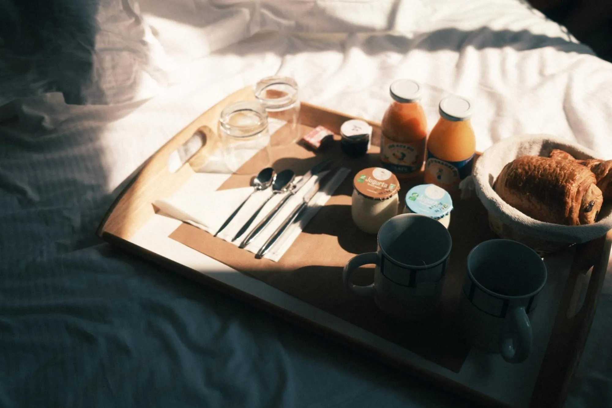 Un plateau de petit-déjeuner avec des viennoiseries, des boissons et de la vaisselle est posé sur un lit.