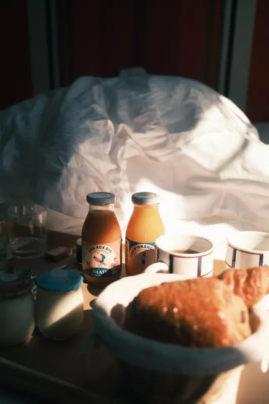 Un petit-déjeuner est servi sur un lit défait, avec des jus d'orange, un croissant et des boissons chaudes.
