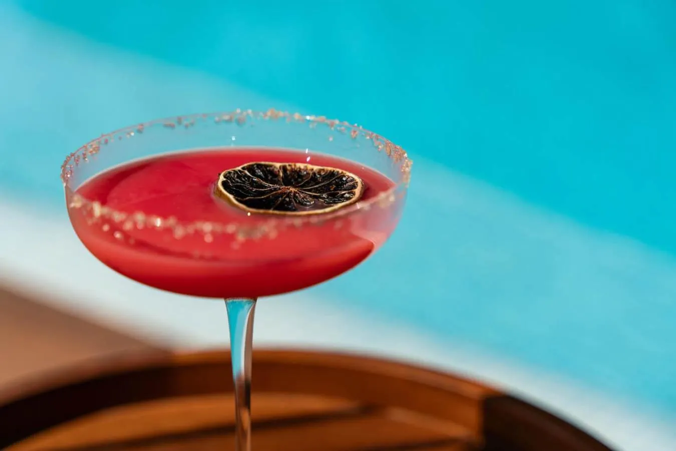 Un cocktail rouge vif dans un verre décoré, posé sur une surface en bois près d'une piscine.
