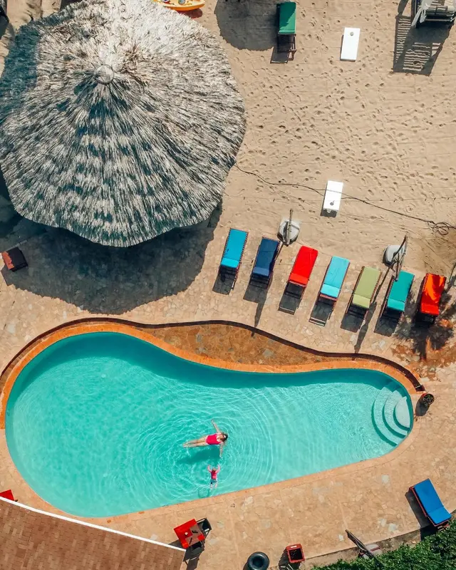 Transats colorés alignés sur une plage de sable fin, avec une piscine turquoise et des parasols en chaume.