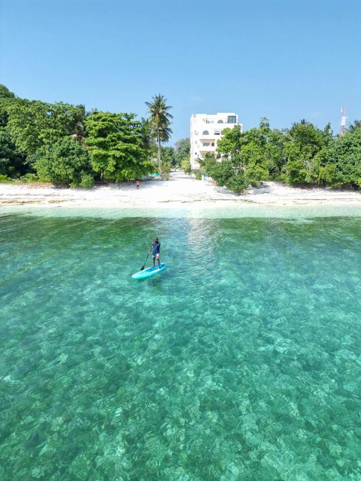 Une personne pratique le stand-up paddle dans une eau turquoise peu profonde près d'une plage de sable blanc.