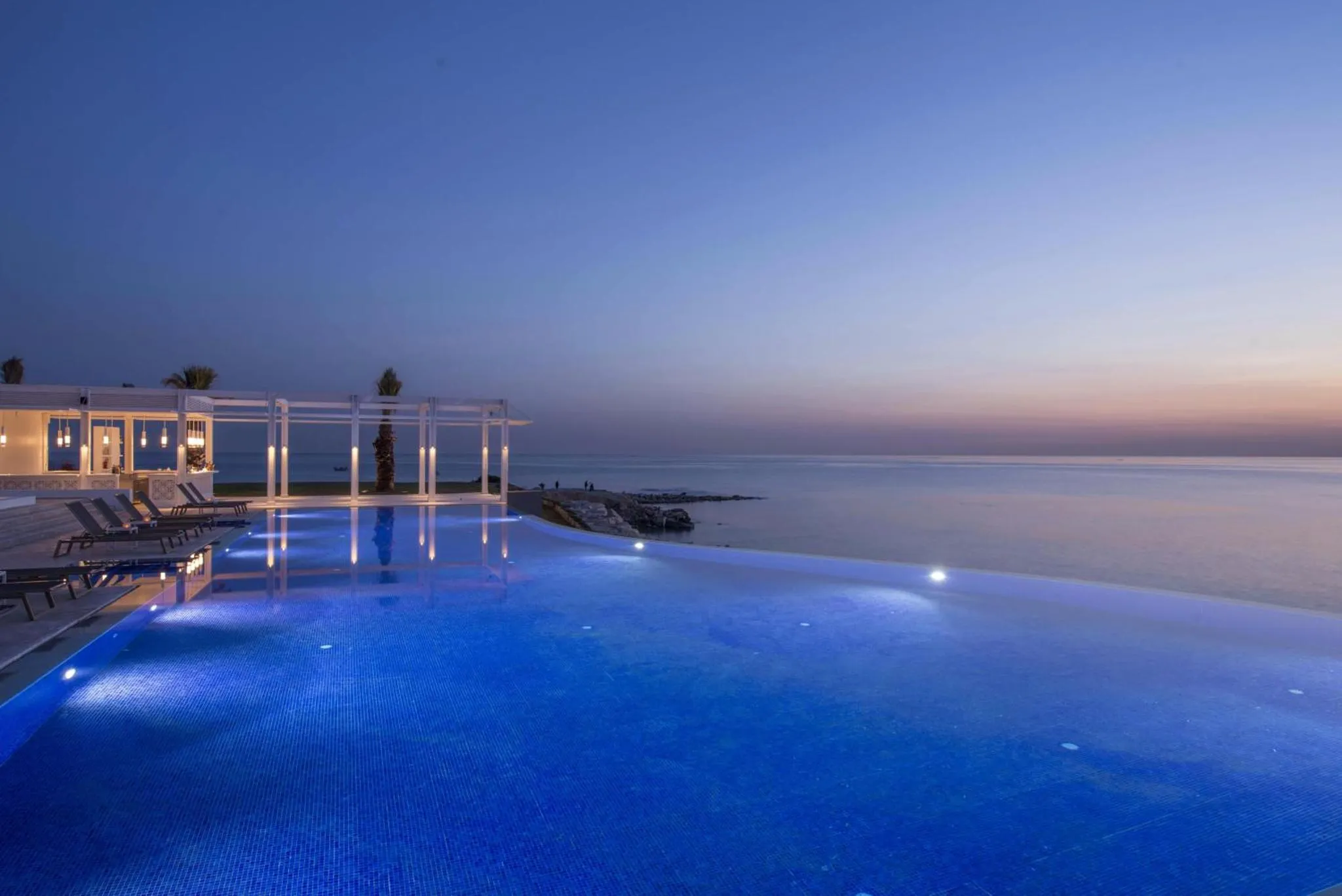 Piscine à débordement illuminée en bleu au crépuscule avec vue sur la mer.