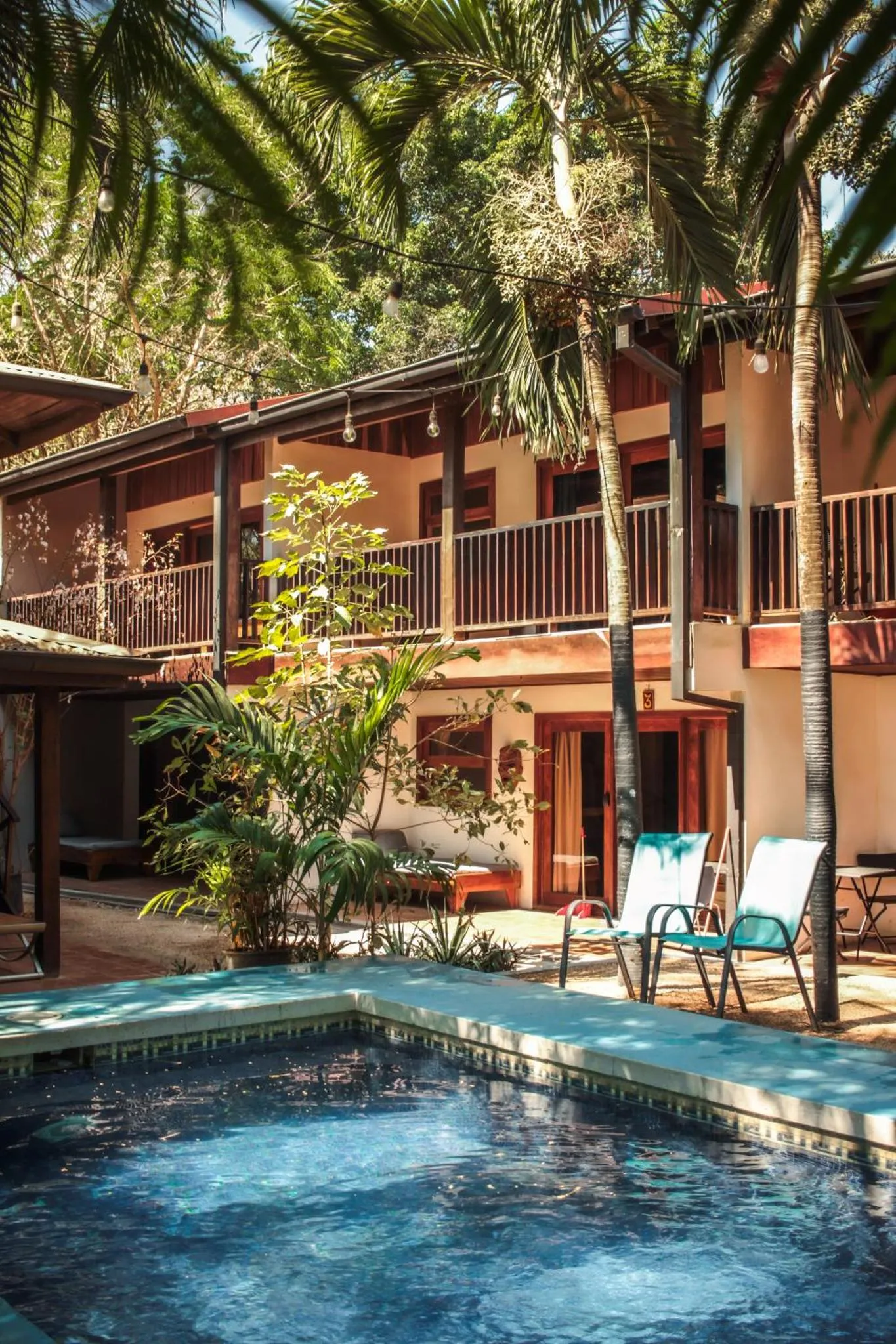 Bâtiment beige à balcons, entouré de végétation tropicale et surplombant une piscine.