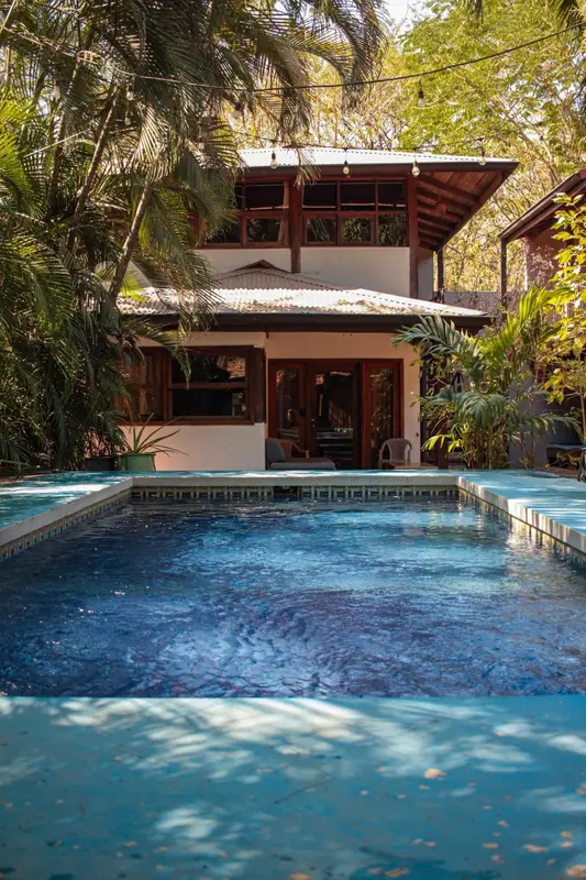 Une piscine rectangulaire devant un bâtiment à deux étages entouré de végétation tropicale.