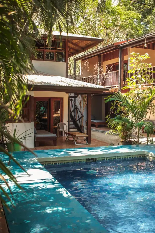Une piscine turquoise au premier plan, avec un bâtiment à l'arrière-plan entouré de végétation tropicale.