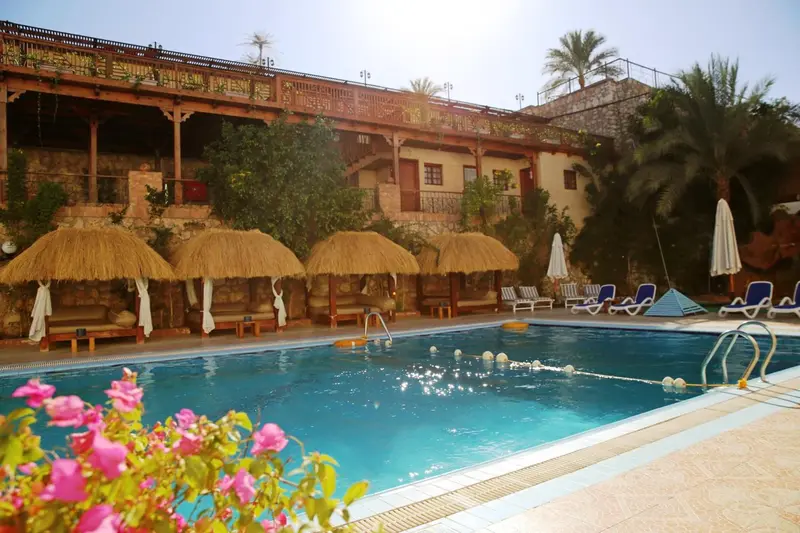 Piscine turquoise bordée de chaises longues et de parasols en chaume devant un bâtiment beige.